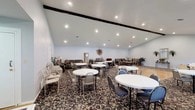 This versatile single-floor gathering space is designed to accommodate a variety of social functions and events. The room is defined by its striking vaulted ceiling with exposed dark beam accents, which adds architectural character and creates an open, airy atmosphere throughout. Recessed lighting is installed across the ceiling, providing consistent, well-distributed illumination.

The flooring presents a deliberate combination of materials: a central hardwood floor section offers a smooth, polished surface ideal for dancing or open activities, while richly patterned floral carpeting surrounds it, delineating the seating and lounge areas. Multiple round tables paired with folding and decorative chairs are arranged throughout the carpeted sections, providing ample seating for gatherings of various sizes.

A row of large windows along one wall invites generous natural light into the space while offering views to the outdoors. Along the perimeter, casual lounge seating including sofas and armchairs creates relaxed conversation areas. Decorative touches such as wall mirrors, potted plants, floor lamps, and wall art contribute a welcoming, homey ambiance. Additional folding chairs and tables are available along the walls, allowing for flexible reconfiguration to suit different event needs.