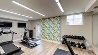 This fitness center is a thoughtfully designed, single-level space built to support a well-rounded workout experience. The room features light wood-style flooring throughout, providing a clean and modern aesthetic that is both practical and visually appealing. Bright linear LED lighting runs across the ceiling, ensuring the entire space is evenly and generously illuminated.

A standout design element is the bold, large-scale geometric mural on the main accent wall, rendered in shades of green, yellow, grey, and white. This eye-catching feature adds energy and personality to the space. Natural light filters in through windows on the exterior wall, complementing the overhead lighting.

The cardio section is equipped with a treadmill, an elliptical trainer, and a rowing machine, each stationed on protective floor mats. A wall-mounted flat-screen television is positioned nearby for entertainment during workouts. A dedicated free weights area includes a fully stocked dumbbell rack, an adjustable weight bench, and a rack of kettlebells.

A floor-to-ceiling mirror along one wall visually expands the space and allows users to monitor their form. The functional training zone includes stability balls, resistance bands, yoga mats, and foam rollers neatly organized on storage racks. A water dispenser and waste receptacle are conveniently located near the exit corridor, adding to the overall functionality of this well-appointed fitness facility.