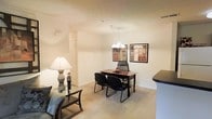 This single-floor residence offers a welcoming and functional layout with a cohesive, neutral color palette throughout. The open-concept living and dining area serves as the heart of the space, seamlessly connecting the two zones for effortless everyday living. The dining area is anchored by a warm wood-toned desk-style table paired with black chairs and illuminated by an elegant multi-arm chandelier overhead, adding a refined touch to the room. Framed wall art provides visual interest and depth to the space. Plush, light-toned carpeting runs throughout the living and dining areas, contributing to a soft and comfortable ambiance underfoot. The living area is complemented by a classic table lamp that adds warm accent lighting. A partial countertop visible at the edge of the frame hints at a kitchen area nearby, suggesting a practical and connected floor plan. The walls are finished in a clean, light neutral tone that reflects light well and enhances the airy feel of the interior. The overall atmosphere is calm, comfortable, and move-in ready, with a straightforward layout designed to maximize usability across the single level.