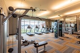 This fitness center in San Antonio, Texas, is a thoughtfully designed, single-floor facility offering a comprehensive range of exercise equipment and functional workout zones. The main workout area features a generous open layout with striped, multi-toned carpet tiles that lend a modern aesthetic throughout the space. Ceiling fans are distributed across the room, ensuring consistent airflow during workouts.

Along one side, a row of treadmills lines a floor-to-ceiling window wall, providing exercisers with outdoor views while maintaining a bright, airy atmosphere. The strength training section is well-appointed with cable machines, a squat rack, adjustable benches, and a dumbbell rack. Large wall-mounted mirrors amplify the sense of space and allow users to monitor their form.

A separate studio area, accessible via sliding barn-style glass doors, houses spinning bikes, stability balls, and additional free weights beneath a dramatic oversized window that floods the room with natural light and views of the surrounding community.

Linear LED lighting fixtures run across the ceiling throughout, providing consistent, even illumination. The overall design balances functionality with a clean, contemporary aesthetic, creating an inviting environment suited to a wide range of fitness activities.