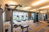 This fitness center in San Antonio, Texas, is a thoughtfully designed, single-floor facility offering a comprehensive range of exercise equipment and functional workout zones. The main workout area features a generous open layout with striped, multi-toned carpet tiles that lend a modern aesthetic throughout the space. Ceiling fans are distributed across the room, ensuring consistent airflow during workouts.

Along one side, a row of treadmills lines a floor-to-ceiling window wall, providing exercisers with outdoor views while maintaining a bright, airy atmosphere. The strength training section is well-appointed with cable machines, a squat rack, adjustable benches, and a dumbbell rack. Large wall-mounted mirrors amplify the sense of space and allow users to monitor their form.

A separate studio area, accessible via sliding barn-style glass doors, houses spinning bikes, stability balls, and additional free weights beneath a dramatic oversized window that floods the room with natural light and views of the surrounding community.

Linear LED lighting fixtures run across the ceiling throughout, providing consistent, even illumination. The overall design balances functionality with a clean, contemporary aesthetic, creating an inviting environment suited to a wide range of fitness activities.