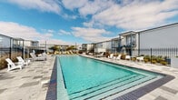 Located at 1500 Lorene Dr in Harvey, Louisiana, this outdoor amenity space centers around a generously sized rectangular swimming pool with clear, vibrant turquoise water and defined lap lanes. The pool deck is finished in a patterned tile surround that provides ample space for lounging and socializing, and is furnished throughout with white Adirondack-style chairs, lounge chairs, and small side tables arranged to take full advantage of the open-air environment. A covered station with a navy blue canopy anchors one end of the pool area, providing shade and housing safety equipment including a life ring. A second shaded section on the opposite side features bright yellow umbrellas and additional seating, adding a pop of color to the space. The entire area is enclosed by a black iron perimeter fence, ensuring safety and privacy. Manicured ornamental plantings and grasses line the fence interior, contributing texture and greenery. The surrounding residential buildings, finished in gray siding with navy blue accents, balconies, and exterior staircases, frame the pool court and create a cohesive community aesthetic. The space benefits from abundant natural light under open Louisiana skies, making it an inviting outdoor retreat.
