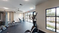 This single-floor fitness center is designed to support a well-rounded workout experience. The space is thoughtfully laid out, with distinct zones accommodating both cardiovascular and strength training activities. The flooring throughout is a durable dark rubber material, well-suited for high-traffic exercise use.

The cardio section is generously outfitted with multiple Spirit-brand treadmills, an elliptical trainer, and a recumbent stationary bike, providing users with a variety of low- and high-impact options. A dedicated alcove area houses additional bikes and offers a quieter corner for focused training. The strength training area features a multi-station cable machine alongside weight benches, supporting a range of resistance exercises.

Large floor-to-ceiling windows along one wall allow ample natural light to fill the space and provide views of the surrounding outdoor environment, creating an open and energizing atmosphere. Ceiling fans and flush-mount light fixtures ensure the room remains well-ventilated and evenly lit. The neutral, light-toned walls contribute to a clean, modern aesthetic throughout.

Additional amenities include a fire extinguisher station, posted safety signage, and access to what appears to be an adjoining restroom or utility room, making this a fully functional and user-friendly fitness facility.