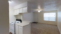 This single-floor space offers a practical and functional layout centered around an open-concept design that connects the kitchen directly to the living area. The kitchen is equipped with white cabinetry, an electric range, an overhead exhaust hood, and a peninsula countertop that serves as a natural divider between the cooking and living spaces. Ample upper cabinet storage keeps the kitchen organized and efficient. The living area features wall-to-wall carpet flooring, lending warmth underfoot, and is illuminated by a ceiling fan with an integrated light fixture. A large window along the far wall allows natural light to fill the room, contributing to a bright and open atmosphere. A textured ceiling runs throughout the space, adding a subtle visual element to the interior. The entry door is conveniently accessible from the living area. The overall aesthetic is clean and neutral, with white walls and light-toned finishes that create a versatile backdrop suitable for a variety of furnishing styles. The seamless flow between the kitchen and living area makes the space feel cohesive and easy to navigate, ideal for everyday living.