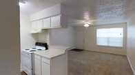 This single-floor space offers a practical and functional layout centered around an open-concept design that connects the kitchen directly to the living area. The kitchen is equipped with white cabinetry, an electric range, an overhead exhaust hood, and a peninsula countertop that serves as a natural divider between the cooking and living spaces. Ample upper cabinet storage keeps the kitchen organized and efficient. The living area features wall-to-wall carpet flooring, lending warmth underfoot, and is illuminated by a ceiling fan with an integrated light fixture. A large window along the far wall allows natural light to fill the room, contributing to a bright and open atmosphere. A textured ceiling runs throughout the space, adding a subtle visual element to the interior. The entry door is conveniently accessible from the living area. The overall aesthetic is clean and neutral, with white walls and light-toned finishes that create a versatile backdrop suitable for a variety of furnishing styles. The seamless flow between the kitchen and living area makes the space feel cohesive and easy to navigate, ideal for everyday living.