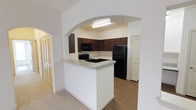 This single-story space showcases a thoughtfully designed kitchen as its centerpiece, framed by an elegant arched entryway that creates a visually striking transition between living areas. The kitchen features rich, dark espresso-toned cabinetry that extends to the upper walls, providing generous storage space. Light granite-style countertops offer a crisp contrast against the darker cabinetry, while a peninsula counter extends outward, creating an ideal space for casual dining or meal preparation. Black appliances, including a refrigerator, range, and over-the-range microwave, lend a cohesive, modern aesthetic throughout the kitchen. The flooring within the kitchen is a warm wood-tone laminate, which transitions to carpet in the adjacent living areas. Neutral wall tones throughout the space contribute to a clean, bright atmosphere. A louvered bi-fold closet door is visible in the adjacent area, hinting at practical storage solutions. Overhead fluorescent lighting ensures the kitchen is well-lit. The open layout and arched architectural detail give the space a welcoming, connected feel.