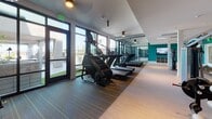 This well-appointed fitness center in San Diego's 92116 neighborhood offers a comprehensive, single-level workout facility designed for a wide range of training styles. The space is divided into distinct functional zones, each thoughtfully equipped and finished.

The cardio area lines the perimeter of expansive floor-to-ceiling glass walls, flooding the room with natural light and providing views of an adjacent outdoor turf area with seating. Treadmills, StairMaster steppers, and elliptical trainers are arranged along this window-facing wall, creating an open, airy workout environment. A large athletic-themed mural adds a dynamic visual element to this section.

The strength and functional training zone features a robust selection of equipment, including cable machines, a multi-functional power rack, TRX suspension trainers, stability balls, and a full dumbbell rack. Floor-to-ceiling mirrors amplify the sense of space and support proper form during training. The durable wood-plank style flooring runs throughout, providing both aesthetic continuity and practical resilience.

A dedicated studio room offers a clean, open floor plan with light hardwood flooring, full-length mirrors, a dramatic black-and-white surf-themed mural, and a wall-mounted TRX training zone with foam rollers and accessories. Recessed and track lighting throughout the facility ensures well-lit conditions at all hours.