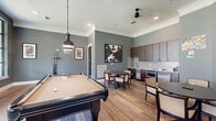 Located at 7600 S Mason Rd in Richmond, TX, this single-floor community lounge and game room is a well-appointed social space designed for recreation and gathering. The open layout accommodates a full-size billiards table as its centerpiece, positioned beneath an industrial-style pendant light that anchors the room's modern aesthetic.

The walls are finished in a cool slate-gray tone, complemented by white crown molding and baseboards that add a clean, polished contrast. Warm hardwood-style flooring runs throughout, providing a cohesive and inviting foundation. Curated artwork, including a bold black-and-white portrait and framed playing card prints, add personality and visual interest to the space.

The kitchenette is outfitted with rich dark wood cabinetry, light granite countertops, a white subway tile backsplash, a stainless steel sink, a built-in oven, and a compact refrigerator — ideal for hosting and casual entertaining. Adjacent to the kitchen, a round dining table with woven-back chairs offers a comfortable spot for smaller gatherings.

Large, multi-pane windows along one wall flood the space with natural light and provide views of the surrounding community grounds. A ceiling fan, recessed lighting, and glass-paneled doors leading to an adjoining fitness area complete this functional and stylishly designed communal space.