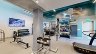 Located at 4150 Jason St in Denver's vibrant LoHi neighborhood, this contemporary two-floor fitness facility delivers a well-rounded workout environment designed for performance and comfort. The lower level serves as the primary strength and functional training zone, featuring exposed concrete ceilings and structural columns that lend an industrial character to the space. A bold teal accent wall adorned with motivational graphics adds visual energy, while large mirrors maximize the sense of depth. This level is equipped with a comprehensive selection of free weights, cable machines, functional trainers, resistance equipment, and rowing machines, complemented by wall-mounted televisions. Large windows and glass entry doors invite natural light and provide views to the surrounding streetscape. A central staircase connects to the upper level, which is designed for cardio and group fitness. The upper floor features warm hardwood flooring, sleek linear pendant lighting, and a mirrored wall alongside a dedicated studio space with glass-paneled doors—ideal for cycling classes or stretching sessions. Treadmills and stair climbers are positioned alongside large windows overlooking the exterior, creating an energizing environment. The open mezzanine railing provides visual connectivity between the two floors, reinforcing the facility's cohesive and modern design throughout.