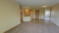 This single-floor residential space offers a practical and functional layout designed for comfortable everyday living. The living area is finished with plush wall-to-wall carpeting, creating a warm and inviting atmosphere, and flows seamlessly into the kitchen and entryway areas.

The kitchen is positioned toward the center of the space and is defined by a half-wall breakfast bar that separates it from the main living area. It features white cabinetry offering generous storage, a double sink, a refrigerator, and a dishwasher, all set on easy-to-clean hard flooring. The kitchen's open connection to the living space promotes an airy, connected feel throughout.

Near the main entry door, a dedicated closet provides convenient storage for coats and everyday essentials. The entry area transitions smoothly into the open living space, giving the layout a logical and welcoming flow. The neutral, light-toned walls throughout the unit reflect ambient light and contribute to the bright, clean aesthetic of the interior.

Overall, this space presents a straightforward and well-organized single-level layout, with defined zones for living, cooking, and entry that make it both practical and livable.