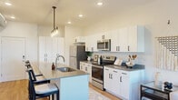 This well-designed 1-bedroom, 1-bathroom unit offers a thoughtful open-concept layout that maximizes every square foot. The kitchen is a standout feature, showcasing crisp white cabinetry with matte black hardware, grey quartz countertops, a full suite of stainless steel appliances, and a spacious center island with bar seating illuminated by contemporary pendant lighting. The kitchen flows seamlessly into a combined dining and living area finished with warm hardwood-style flooring. Exposed ductwork adds an industrial-modern edge, while large windows with floor-to-ceiling drapes flood the space with natural light and offer views of the surrounding neighborhood. A private balcony door in the living area further extends the connection to the outdoors. The bedroom is a comfortable retreat featuring a generous window with outdoor views, a ceiling fan, and soft carpeting. The full bathroom is clean and modern, featuring white cabinetry with grey countertops, a tub-shower combination, and hardwood-style flooring. A walk-through closet connects the bathroom to the bedroom, providing convenient and organized storage. Recessed lighting throughout maintains a bright, contemporary ambiance across the entire unit.