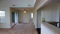 This single-story space showcases an open, flowing layout that connects the main living area to additional rooms beyond. The living room is finished with neutral-toned carpet and features a ceiling fan with an integrated light fixture, providing both comfort and illumination. A sliding glass door along one wall invites natural light into the space while offering convenient access to an outdoor area, where a tiled patio or balcony is partially visible.

A wide passageway leads from the living area into a hallway that connects to further rooms, one of which receives natural light through a window, hinting at a bright and airy atmosphere throughout. The walls are finished in a warm, neutral tone that complements the soft carpeting underfoot.

To one side, a raised granite-topped breakfast bar forms a natural divider between the living space and the kitchen, offering a casual dining or serving surface. The kitchen area, partially visible from this vantage point, includes a mirrored or reflective wall element. HVAC vents are positioned in the ceiling, ensuring climate control throughout. Overall, the space presents a practical and comfortable open-concept design with functional, everyday amenities.