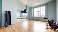 Located at 2525 Sycamore Farms Dr NW in Concord, NC, this well-appointed fitness studio offers a clean, purpose-built environment designed to support a variety of workout routines. The space is finished with smooth light hardwood flooring throughout, providing an ideal surface for both cardio and low-impact exercise.

One wall features a large floor-to-ceiling mirror panel paired with a wall-mounted ballet barre, making the studio well-suited for barre classes, stretching, and dance-inspired workouts. A ceiling-mounted flat-screen television is centrally positioned, allowing for instructor-led video workouts or fitness programming visible from anywhere in the room.

A row of spin bikes is stationed along one side of the studio, offering a dedicated cycling area, while a freestanding medicine ball rack provides easy access to weighted fitness equipment. The studio is bathed in natural light through large, multi-pane windows that frame views of the surrounding neighborhood and open sky, contributing to an energizing and open atmosphere.

The walls are painted in a soft, cool blue-gray tone that gives the space a calm and focused ambiance. High ceilings enhance the sense of spaciousness, while recessed and pendant linear lighting ensures the room is well-lit at all times. A glass partition wall connects the studio to an adjacent fitness area, allowing visual continuity between spaces.