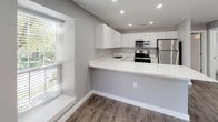 This single-floor space showcases a thoughtfully designed, contemporary interior with a cohesive neutral palette of cool grays and crisp whites. The kitchen is a standout feature, equipped with flat-front white cabinetry extending to the ceiling for ample storage, bright white quartz countertops, and a full suite of stainless steel appliances including a refrigerator, range, microwave, and dishwasher. A peninsula-style counter extends into the living area, creating a natural divide between the kitchen and main living space while offering additional prep and seating space. A window positioned near the kitchen allows natural light to filter in, enhancing the open feel. Recessed lighting throughout ensures consistent illumination.

The bedroom is well-proportioned, featuring wood-look flooring, soft gray walls, a ceiling fan with integrated lighting, and a window dressed with horizontal blinds. The bathroom maintains the clean aesthetic with a tiled tub-surround, built-in niche shelving, chrome fixtures, and tile flooring. Consistent wood-look flooring runs throughout the main living areas, tying the space together with a warm yet modern character.