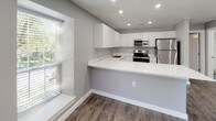 This single-floor space showcases a thoughtfully designed, contemporary interior with a cohesive neutral palette of cool grays and crisp whites. The kitchen is a standout feature, equipped with flat-front white cabinetry extending to the ceiling for ample storage, bright white quartz countertops, and a full suite of stainless steel appliances including a refrigerator, range, microwave, and dishwasher. A peninsula-style counter extends into the living area, creating a natural divide between the kitchen and main living space while offering additional prep and seating space. A window positioned near the kitchen allows natural light to filter in, enhancing the open feel. Recessed lighting throughout ensures consistent illumination.

The bedroom is well-proportioned, featuring wood-look flooring, soft gray walls, a ceiling fan with integrated lighting, and a window dressed with horizontal blinds. The bathroom maintains the clean aesthetic with a tiled tub-surround, built-in niche shelving, chrome fixtures, and tile flooring. Consistent wood-look flooring runs throughout the main living areas, tying the space together with a warm yet modern character.