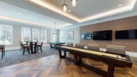 This single-floor entertainment and game lounge is thoughtfully designed to offer a diverse range of recreational activities within a cohesive, stylish environment. The space is anchored by elegant herringbone-patterned hardwood flooring that transitions seamlessly into plush, patterned carpeting, defining distinct activity zones throughout. Tray ceilings with integrated ambient LED lighting add architectural depth and a warm glow to the entire room, complemented by decorative pendant fixtures that enhance the refined atmosphere.

A dedicated lounge area features a row of plush, light-toned leather recliner chairs positioned in front of a large wall-mounted flat-screen television set against a warm wood plank accent wall — ideal for viewing and relaxation. Adjacent to this, a full-size shuffleboard table is flanked by additional seating chairs, with dual flat-screen monitors mounted on the accent wall for added entertainment.

The billiards zone offers a regulation pool table set beneath abundant natural light from large windows, accompanied by high-top pub tables and bar stools for casual socializing. A dedicated arcade section houses electronic dartboards, a multi-game arcade cabinet, and a classic pinball machine. The overall ambiance is sophisticated yet inviting, balancing modern amenities with warm, upscale finishes throughout.