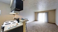 This single-floor residential unit offers a functional and comfortable open-concept layout. The kitchen and living areas flow seamlessly into one another, creating a connected and practical living space. The kitchen is equipped with an electric range, an overhead range hood, and a breakfast bar with bar seating — ideal for casual dining. Dark cabinetry above the range contrasts with the lighter tones throughout the rest of the space.

The living area is finished with wall-to-wall carpeting, providing a warm and comfortable feel underfoot. A textured ceiling adds a subtle visual element throughout the unit. Neutral-toned walls keep the space feeling open and versatile.

A prominent sliding glass door fitted with chevron-patterned vertical blinds serves as a focal point of the living area, allowing an abundance of natural light to fill the room while also offering access to an outdoor space. The overall ambiance of the unit is cozy and practical, suited for everyday living. The open layout and shared natural light between the kitchen and living room contribute to a welcoming and airy atmosphere throughout the home.