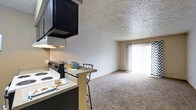 This single-floor residential unit offers a functional and comfortable open-concept layout. The kitchen and living areas flow seamlessly into one another, creating a connected and practical living space. The kitchen is equipped with an electric range, an overhead range hood, and a breakfast bar with bar seating — ideal for casual dining. Dark cabinetry above the range contrasts with the lighter tones throughout the rest of the space.

The living area is finished with wall-to-wall carpeting, providing a warm and comfortable feel underfoot. A textured ceiling adds a subtle visual element throughout the unit. Neutral-toned walls keep the space feeling open and versatile.

A prominent sliding glass door fitted with chevron-patterned vertical blinds serves as a focal point of the living area, allowing an abundance of natural light to fill the room while also offering access to an outdoor space. The overall ambiance of the unit is cozy and practical, suited for everyday living. The open layout and shared natural light between the kitchen and living room contribute to a welcoming and airy atmosphere throughout the home.
