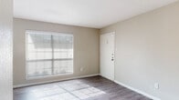 This single-story residence presents a clean, neutral aesthetic throughout, with warm beige walls and white trim creating a cohesive and inviting atmosphere. The main living area is finished with durable wood-look flooring and benefits from generous natural light streaming through a large window fitted with horizontal blinds, casting bright patterns across the floor. A dedicated entry door with secure hardware opens directly into this welcoming space.

The kitchen is thoughtfully appointed with dark cabinetry, granite countertops, and black appliances including a refrigerator and range. An adjacent open shelving nook provides convenient storage and connects the kitchen to the rest of the living areas via a smoothly flowing layout.

A separate room features plush carpeting and a ceiling fan with integrated lighting, along with a sliding glass door flanked by vertical blinds that opens to an outdoor area. This carpeted space also connects to additional rooms, including a well-sized bedroom with its own ceiling fan and built-in closet access.

The bathroom is finished with a dark wood vanity topped with granite, a large mirror, and a stylish light fixture overhead. An additional flexible space with wood-look flooring includes a pass-through niche with accent lighting, offering functional versatility throughout the unit.