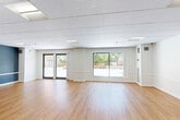 Located at 36 Dartmouth St in Malden, MA, this single-floor commercial space offers a well-designed, open layout ideal for a variety of uses. The main area is expansive and bright, featuring warm hardwood-style flooring throughout and a clean white color palette accented by slate-blue walls in select areas, creating a modern yet welcoming atmosphere. A drop tile ceiling with recessed fluorescent lighting ensures consistent, even illumination across the entire floor.

Large sliding windows and a glass sliding door along one wall flood the main area with natural light while offering views of the outdoor surroundings. Structural columns are neatly finished in white, adding architectural character without disrupting the open flow of the space. White chair rail molding lines the lower portion of the walls, lending a polished, professional appearance.

The kitchen is fully equipped with white shaker-style cabinetry, light countertops, a gas cooktop, a stainless steel dishwasher, a full-size stainless steel refrigerator, and a built-in range hood. The cabinetry features a bold navy accent wall above, adding a contemporary design touch. A pass-through window connects the kitchen to the main area, making it well-suited for serving or collaborative use. This space is thoughtfully designed to support a range of professional or community-oriented functions.