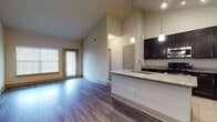 This single-floor residence is defined by its striking vaulted ceilings, which lend an airy, open quality to the main living and kitchen areas. Rich hardwood-style flooring runs throughout the living spaces, complementing the neutral warm-toned walls and crisp white baseboards.

The kitchen is a functional centerpiece, featuring dark espresso cabinetry that extends toward the ceiling, granite countertops, and a large peninsula island with an undermount sink and pendant lighting above. Stainless steel appliances—including a refrigerator, microwave, and range—add a modern touch, while tile flooring grounds the cooking area. Track lighting provides additional illumination overhead.

The open layout connects the kitchen seamlessly to the living area, which benefits from natural light through a large windowed entry door and nearby windows. The bedroom is a well-proportioned space with a ceiling fan, an arched window overlooking greenery, and access to a spacious walk-in closet with built-in shelving.

The bathroom includes a glass-enclosed walk-in shower with full tile surround and built-in shelving, paired with granite countertop finishes consistent throughout the unit. The overall ambiance is clean, contemporary, and move-in ready.