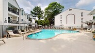 Located in the Virginia-Highland neighborhood of Atlanta, GA, this outdoor patio and pool area offers a well-appointed amenity space designed for relaxation and recreation. The centerpiece is a generously sized, curved resort-style swimming pool with crystal-clear blue water, surrounded by a broad concrete deck that provides ample room for lounging. Rows of chaise lounge chairs and striped umbrellas line the pool's perimeter, offering both sun and shade options throughout the day. A charming poolside cabana structure features arched openings, ceiling fans, and additional seating, providing a shaded retreat with an inviting, classic aesthetic. The surrounding architecture reflects a traditional Southern style, with white-painted brick exteriors accented by distinctive brown brick arched detailing. Raised brick planters filled with vibrant red seasonal flowers add a decorative touch near the building entry. A dedicated outdoor dining area equipped with slatted wood tables, lime green chairs, and bold black-and-white striped umbrellas creates an inviting space for al fresco dining. Mature trees frame the perimeter, offering natural shade and a lush green backdrop. The entire area is enclosed by a sleek metal fence, ensuring privacy while maintaining an open, airy feel throughout the space.