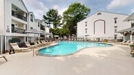 Located in the Virginia-Highland neighborhood of Atlanta, GA, this outdoor patio and pool area offers a well-appointed amenity space designed for relaxation and recreation. The centerpiece is a generously sized, curved resort-style swimming pool with crystal-clear blue water, surrounded by a broad concrete deck that provides ample room for lounging. Rows of chaise lounge chairs and striped umbrellas line the pool's perimeter, offering both sun and shade options throughout the day. A charming poolside cabana structure features arched openings, ceiling fans, and additional seating, providing a shaded retreat with an inviting, classic aesthetic. The surrounding architecture reflects a traditional Southern style, with white-painted brick exteriors accented by distinctive brown brick arched detailing. Raised brick planters filled with vibrant red seasonal flowers add a decorative touch near the building entry. A dedicated outdoor dining area equipped with slatted wood tables, lime green chairs, and bold black-and-white striped umbrellas creates an inviting space for al fresco dining. Mature trees frame the perimeter, offering natural shade and a lush green backdrop. The entire area is enclosed by a sleek metal fence, ensuring privacy while maintaining an open, airy feel throughout the space.