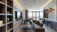 This elegantly designed single-floor high-rise space offers a seamless blend of modern luxury and functional living. The main living area is anchored by a large wall-mounted television set against a textured white panel feature wall, complemented by warm cove lighting that runs along the ceiling perimeter, creating a refined ambiance throughout. A built-in open shelving unit in light wood tones provides generous display storage, adding both character and practicality to the space.

The lounge is furnished with plush, earth-toned sectional sofas arranged around a pair of sleek metal-framed coffee tables, all resting on a patterned area rug over large-format tile flooring. A striking marble-topped bar counter with black bar stools defines a dedicated entertaining zone, dramatically lit by a cluster of sculptural copper pendant lights that serve as a standout design feature.

Floor-to-ceiling glass doors and expansive windows flood the interior with natural light while framing sweeping panoramic city views. These doors open directly onto a private balcony furnished with outdoor seating, extending the living experience outdoors. A compact wet bar area with built-in appliances and cabinetry provides convenient in-unit amenities. Bold contemporary artwork adorns the walls, completing the space's sophisticated, curated aesthetic.