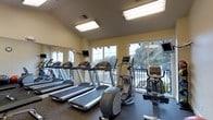 This fitness center is thoughtfully designed to provide a motivating and functional workout environment. The space is outfitted with a range of cardio equipment, including multiple treadmills, elliptical trainers, and a stationary bike, catering to a variety of exercise preferences and fitness levels. Large windows line the walls, flooding the room with abundant natural light and offering views of the surrounding outdoor landscape, creating an open and energizing atmosphere. The light yellow walls and flush-mounted ceiling fixtures contribute to a bright, clean interior aesthetic. Mounted televisions are positioned throughout the space for entertainment during workouts. A full-length mirror along one wall enhances the sense of space and allows users to monitor their form. The flooring features durable, rubber-textured material well-suited for fitness use. The single-floor layout ensures easy accessibility, and the overall design balances practicality with comfort, making it a well-rounded facility for daily exercise routines.