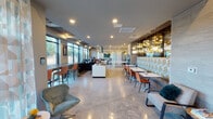 This thoughtfully designed single-floor commercial space, identified as The Cooper, combines hospitality and professional functionality in a cohesive, modern aesthetic. The main café and dining area is an open, airy space anchored by polished concrete floors that extend throughout the entire layout. A central white service island serves as the focal point, flanked by a full coffee and beverage station. Along one wall, banquette seating features decorative patterned wallcovering beneath a striking row of amber glass pendant lights, creating a warm contrast to the otherwise cool-toned palette. Large floor-to-ceiling windows along the perimeter flood the space with natural light and offer views of surrounding greenery.

Transitioning deeper into the space, a lounge area near the entrance features eclectic mid-century-inspired seating in teal, blue, and rust tones set against bold yellow accent walls and curated artwork. A branded entry mat and glass-paneled doors mark the formal arrival point.

Further in, a private meeting and workspace zone features glass-walled conference rooms fitted with sleek office chairs and whiteboards. Sculptural multi-arm pendant lighting adds an artistic touch overhead, while a central white worktable provides collaborative space between rooms. The overall ambiance balances a welcoming café atmosphere with polished, professional functionality.