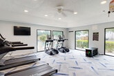 This single-level fitness center is designed to support a comprehensive workout experience within a spacious, open layout. The room is finished with light gray walls and a textured ceiling, lending a clean and modern aesthetic throughout. Recessed lighting is distributed evenly across the ceiling and is complemented by a ceiling fan, ensuring the space remains well-lit and ventilated during use.

The cardio zone is anchored by multiple True-brand treadmills and elliptical machines, each equipped with individual display screens. A wall-mounted flat-screen television provides additional entertainment for users during extended sessions. A multi-functional cable trainer occupies a dedicated area of the room, alongside a vertical medicine ball rack stocked with weighted balls in varying sizes, supporting strength and functional training routines.

Large, black-framed windows and sliding glass doors line the exterior walls, flooding the interior with natural light and offering views of the surrounding landscaped grounds. The flooring features a distinctive patterned carpet with blue and gray geometric accents, contributing to the room's polished appearance. An open stretch area near the windows provides additional space for floor exercises or cool-down routines.