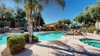 This outdoor amenity space centers around a generously sized freeform swimming pool with clear turquoise water, designed to evoke a resort-like atmosphere. The pool's curving shape adds visual interest and flows naturally into the surrounding hardscape, which features a broad, textured concrete deck well-suited for lounging and socializing. A separate spa or wading area is positioned adjacent to the main pool, accented by decorative boulders that contribute to the desert-inspired aesthetic.

A covered gazebo provides a shaded retreat along the pool's edge, offering a comfortable outdoor gathering spot during warm, sunny days. The sundeck is furnished with multiple lounge chairs, ideal for relaxing in the abundant natural sunlight characteristic of the Southwest. Wrought iron fencing encloses the entire pool area, ensuring both security and an open sightline to the surrounding landscaping.

The grounds are thoughtfully landscaped with tall mature palm trees, desert shrubs, flowering plants, and native greenery that provide shade and visual texture. The multi-story residential buildings visible in the background feature a Southwestern architectural style with warm earth tones, tile rooflines, and stucco exteriors, lending a cohesive desert community ambiance to the entire outdoor space.