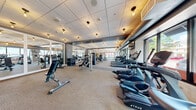 Located at 430 E Walnut St in Springfield, Missouri, this fitness center offers a thoughtfully designed exercise space that accommodates a wide range of workout needs. The single-floor layout provides generous room for movement and equipment, creating an open, uncluttered environment for users.

The exercise room is outfitted with a full lineup of True-brand cardio equipment, including treadmills and elliptical machines, arranged along a wall of large windows that offer views of the surrounding streetscape and flood the space with natural light. Strength training is well represented through multiple cable and resistance machines, adjustable benches, and a dedicated free weights area with a tiered dumbbell rack.

Large framed mirrors are strategically positioned throughout the room, visually expanding the space and allowing users to monitor their form. The flooring consists of neutral, low-pile carpet suited for both machine use and floor exercises. Overhead, a drop tile ceiling is fitted with warm Edison-style pendant lights that complement the modern, clean aesthetic of the room.

Exposed ductwork near the cardio section adds an industrial touch, while the overall design palette of gray and white keeps the atmosphere modern and professional. The space also includes a beverage vending unit for post-workout refreshment. Altogether, this fitness center delivers a fully equipped, well-lit, and welcoming environment for a complete workout experience.
