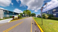 Located at 5903 Northwest 57th Court in Tamarac, Florida, this space serves as the leasing office entry point for the Osprey residential community. The exterior approach sets a welcoming tone, with a well-maintained access road flanked by lush, manicured green lawns, mature shade trees, and tropical palms — all characteristic of South Florida's vibrant landscape. The community's buildings, finished in a distinctive teal and gray color scheme, are visible from the entrance alongside professionally landscaped grounds featuring colorful plantings and yellow-curbed roadways that guide visitors through the property. A clearly marked leasing office sign greets prospective residents at the entrance. The space itself is compact and focused in function, designed to serve as a practical point of contact within the broader community setting. The surrounding outdoor environment is bright and open, benefiting from Florida's abundant natural light beneath expansive skies. The overall ambiance of the entry and leasing area reflects a community that prioritizes curb appeal and accessibility, offering a polished first impression to all who arrive at this Tamarac address.
