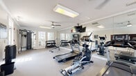 Located in Spanish Fort, Alabama, this fitness center offers a well-equipped, single-floor exercise space designed to support a comprehensive workout routine. The room features a clean, neutral color palette with light gray carpeted flooring, white trim, and crown molding, lending a polished and inviting atmosphere.

The exercise room is outfitted with a diverse selection of equipment, including Spirit-brand treadmills, an elliptical trainer, a stationary bike, and a rowing machine for cardio training. A multi-function cable machine and an adjustable weight bench serve strength training needs, while a tiered dumbbell rack stocked with a full range of free weights provides additional versatility. Exercise balls are also available for functional fitness routines.

Full-length mirrored walls span one side of the room, visually expanding the space and allowing users to monitor their form during workouts. Multiple ceiling fans ensure comfortable air circulation throughout, and recessed and overhead lighting keep the room bright. Large windows with blinds allow ample natural light to filter in while maintaining privacy.

A wall-mounted flat-screen television adds entertainment options during workouts. Near the entrance, a water cooler, waste receptacle, and posted fitness center rules reflect a thoughtfully maintained and community-oriented facility. A glass-panel door offers easy access and connects the space to the surrounding exterior grounds.