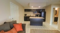 This single-floor space offers a practical and modern open-concept layout that seamlessly connects the kitchen and living areas. The kitchen is a standout feature, outfitted with sleek dark cabinetry that contrasts beautifully against the light-toned walls and white tile flooring. A central kitchen island provides additional counter space, ideal for meal preparation or casual dining. Black appliances, including a refrigerator, range, microwave, and dishwasher, complement the dark cabinetry for a cohesive, contemporary aesthetic. The sink is positioned along the back wall beneath upper cabinets, maximizing storage and workspace efficiency. Recessed ceiling lighting brightens the kitchen, creating a clean and well-lit environment. The living area flows naturally from the kitchen, featuring light tile flooring that extends throughout the open space, accented by a cozy area rug. The living space is highlighted by a comfortable sofa with warm orange accent pillows, lending a welcoming touch to the overall atmosphere. Neutral wall tones keep the space feeling bright and open, while the single-floor layout ensures easy accessibility and a smooth, connected flow throughout the entire unit.