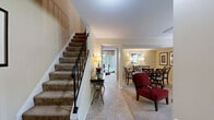 Located at 8800 Howard Hills Dr in Savage, MD, this two-story home offers a thoughtfully organized layout spread across two floors. The main level features a combined dining and living room, a kitchen, and a full bathroom. The dining area is elegantly appointed, with warm neutral walls, decorative framed artwork, and ample space for a full dining table. The adjacent living room is bright and welcoming, featuring carpet flooring, natural light from large windows with views of surrounding greenery, and comfortable proportions. The galley-style kitchen makes efficient use of space, showcasing light maple cabinetry, granite-look countertops, white appliances, and bold red accent walls that lend the room energy and personality. A full bathroom on this level features eye-catching horizontal black and cream striped walls with tile flooring. The upper floor houses both bedrooms, a second full bathroom, and a dedicated office. The spacious primary bedroom includes a closet and soft carpet flooring, while the second bedroom benefits from natural light and treetop views through large windows. The second bathroom offers a tub-shower combination with tile surround. The office provides a quiet, functional retreat with windows overlooking mature trees. Throughout, the home balances practical design with distinctive decorative touches.