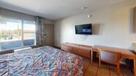 This compact, single-floor unit at 13031 Central Ave NE in Albuquerque, New Mexico offers a practical and self-contained layout comprising one bedroom, one bathroom, and a full kitchen. The bedroom is the largest area of the unit, featuring carpeted flooring, a full-size bed with a Southwestern-patterned bedspread, a wall-mounted flat-screen TV, a low dresser, and a small desk with seating. A window with curtains allows natural light into the room, and a window-unit air conditioner provides climate control. The kitchen is efficiently laid out with warm-toned wood cabinetry, laminate countertops, a stainless steel sink, a built-in cooktop, an over-the-range microwave, and a stainless steel refrigerator. Hardwood-style flooring adds warmth to the kitchen area. The bathroom is straightforward and functional, featuring light tile flooring, a tiled shower-tub combination with a curtain, a toilet, and a towel rack. The overall aesthetic of the unit is simple and utilitarian, catering to those seeking a no-frills, move-in-ready space with essential amenities in a convenient Albuquerque location.