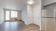 This single-floor unit offers a clean, modern aesthetic throughout its well-designed layout. The entry foyer opens into a bright, open-concept living area featuring light wood-tone luxury vinyl plank flooring, white walls, and track lighting overhead. A glass door at the far end of the living space welcomes natural light and provides outdoor views, enhancing the airy feel of the room.

The kitchen is efficiently designed and fully equipped with stainless steel appliances, including a refrigerator, electric range, and over-the-range microwave. White shaker-style cabinetry extends to the ceiling, paired with dark countertops, a subway tile backsplash, and a stainless steel sink. Track lighting illuminates the workspace effectively.

The bedrooms feature plush wall-to-wall carpeting and neutral toned walls. One bedroom includes a walk-in closet with built-in shelving, while another offers a window with a garden view and a baseboard heater. Both rooms are generously sized with ceiling light fixtures.

The bathroom is finished with light hardwood-style flooring, a full soaking tub with a curved shower rod, white cabinetry, dark countertops, and a large vanity mirror with pendant lighting above. The overall design is cohesive, contemporary, and move-in ready.