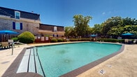 This outdoor pool area presents a well-maintained and inviting recreational space, characterized by a generously sized swimming pool with clear turquoise water and a dark-tiled border that adds a polished, defined edge to the design. Depth markers are visible along the pool's perimeter, and metal entry rails provide safe and convenient access to the water. The pool deck is composed of a smooth concrete surround, offering ample space for lounging and circulation. Surrounding the pool, multiple seating areas are furnished with chairs and tables shaded by vibrant blue umbrellas, creating comfortable spots for relaxation. Lush, mature trees frame the space and provide natural shade, while neatly trimmed hedges and green lawn areas contribute to a well-kept, resort-like ambiance. A prominent classic brick building with large arched windows and French doors anchors one side of the pool area, lending architectural character to the space. On the opposite side, a stone retaining wall with tiered greenery adds depth and a natural boundary. The open sky above floods the entire area with abundant natural light, enhancing the bright and airy atmosphere. The combination of traditional architecture, verdant landscaping, and thoughtfully arranged outdoor furnishings creates a cohesive and appealing community amenity space.