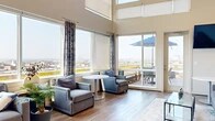Located at 3232 Eastern Ave in Baltimore, MD, this thoughtfully designed lounge space offers an impressive blend of architectural drama and modern comfort. The space is defined by its soaring double-height ceilings and an expansive wall of floor-to-ceiling windows that flood the interior with abundant natural light while framing sweeping panoramic views of the Baltimore cityscape. Warm hardwood-style flooring runs throughout, complementing the neutral tones of the walls and creating a cohesive, inviting atmosphere.

The main lounge area is generously appointed with plush gray sofas, accent armchairs, and a sleek glass coffee table, all arranged to encourage conversation and relaxation. A secondary seating nook features intimate round tables and chairs positioned directly beside the windows and a sliding glass door, which opens onto a furnished outdoor deck — complete with a patio table and umbrella — offering a seamless indoor-outdoor experience with unobstructed city views.

A well-equipped kitchenette serves the space with rich wood cabinetry, light stone countertops, a stainless steel sink, a mosaic tile backsplash, and a wall-mounted television. Decorative sculptural wall art adds a distinctive cultural touch to one of the seating areas, enhancing the overall character of the space. The combination of high design, ample natural light, and elevated outdoor access makes this a versatile and visually striking venue.