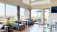 Located at 3232 Eastern Ave in Baltimore, MD, this thoughtfully designed lounge space offers an impressive blend of architectural drama and modern comfort. The space is defined by its soaring double-height ceilings and an expansive wall of floor-to-ceiling windows that flood the interior with abundant natural light while framing sweeping panoramic views of the Baltimore cityscape. Warm hardwood-style flooring runs throughout, complementing the neutral tones of the walls and creating a cohesive, inviting atmosphere.

The main lounge area is generously appointed with plush gray sofas, accent armchairs, and a sleek glass coffee table, all arranged to encourage conversation and relaxation. A secondary seating nook features intimate round tables and chairs positioned directly beside the windows and a sliding glass door, which opens onto a furnished outdoor deck — complete with a patio table and umbrella — offering a seamless indoor-outdoor experience with unobstructed city views.

A well-equipped kitchenette serves the space with rich wood cabinetry, light stone countertops, a stainless steel sink, a mosaic tile backsplash, and a wall-mounted television. Decorative sculptural wall art adds a distinctive cultural touch to one of the seating areas, enhancing the overall character of the space. The combination of high design, ample natural light, and elevated outdoor access makes this a versatile and visually striking venue.