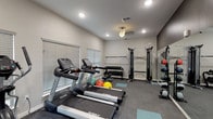 This fitness center is designed to support a comprehensive workout experience within a single, efficiently organized space. The room features large windows fitted with horizontal blinds, allowing natural light to fill the interior while maintaining privacy. Recessed lighting and a ceiling fan overhead ensure the space remains well-lit and comfortable during use. The floors are covered in large-format dark slate-toned tiles, with teal exercise mats positioned throughout to define workout zones.

The cardio area is anchored by multiple Spirit-brand treadmills aligned along one side of the room, accompanied by an elliptical trainer and a recumbent stationary bike, offering a variety of low- and high-impact aerobic options. Medicine balls and stability balls are accessible nearby, supporting functional and core training.

The strength and resistance zone features a multi-cable functional trainer, a wall-mounted barbell rack, a tiered dumbbell rack with a full range of free weights, and a flat weight bench. A large wall mirror is positioned to allow users to monitor their form during exercises. Wall-mounted informational signage provides guidance throughout the space. The overall atmosphere is clean, modern, and practical, providing a focused environment suited for a well-rounded fitness routine.