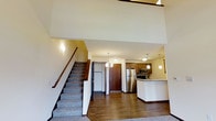 This two-floor residence offers a thoughtfully designed layout that balances open living with functional space across both levels. The main level immediately impresses with its soaring double-height ceiling, which creates a bright, airy atmosphere throughout the open-concept living and kitchen area. Rich hardwood-style flooring runs across the main level, adding warmth and visual continuity to the space.

The kitchen is a standout feature, showcasing medium-toned wood cabinetry, stainless steel appliances including a refrigerator and microwave, and a tile backsplash that adds a contemporary touch. A peninsula with a light-colored countertop provides additional workspace and serves as a natural divider between the kitchen and living areas. Pendant lighting above the peninsula and throughout the kitchen enhances both functionality and ambiance.

A carpeted staircase with a wooden handrail leads to the upper level, where additional living or sleeping spaces are found. Wall sconces along the stairwell provide subtle, warm lighting. The front entry is defined by a solid wood door, offering a welcoming and secure point of arrival. Overall, the space presents a clean, modern aesthetic with quality finishes throughout.