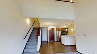 This two-floor residence offers a thoughtfully designed layout that balances open living with functional space across both levels. The main level immediately impresses with its soaring double-height ceiling, which creates a bright, airy atmosphere throughout the open-concept living and kitchen area. Rich hardwood-style flooring runs across the main level, adding warmth and visual continuity to the space.

The kitchen is a standout feature, showcasing medium-toned wood cabinetry, stainless steel appliances including a refrigerator and microwave, and a tile backsplash that adds a contemporary touch. A peninsula with a light-colored countertop provides additional workspace and serves as a natural divider between the kitchen and living areas. Pendant lighting above the peninsula and throughout the kitchen enhances both functionality and ambiance.

A carpeted staircase with a wooden handrail leads to the upper level, where additional living or sleeping spaces are found. Wall sconces along the stairwell provide subtle, warm lighting. The front entry is defined by a solid wood door, offering a welcoming and secure point of arrival. Overall, the space presents a clean, modern aesthetic with quality finishes throughout.