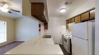 This single-floor space offers a practical and well-organized layout designed to maximize functionality. The kitchen is a standout feature, showcasing a galley-style configuration that makes efficient use of space. Dark wood upper cabinetry lines the walls, providing generous storage, while a light-toned countertop stretches across one side, offering ample workspace for meal preparation. A double-basin sink is neatly integrated into the counter, complemented by overhead storage above. On the opposing wall, white appliances — including an electric coil range with an overhead exhaust hood and a full-size refrigerator — are neatly arranged for a clean, cohesive look. The flooring features dark wood-style material that contrasts with the light walls, lending warmth to the space. Flush ceiling lighting provides consistent, even illumination throughout. A door at the end of the kitchen leads to an adjacent area, suggesting a connected and accessible floor plan. The overall ambiance is clean, neutral, and move-in ready, with a no-frills aesthetic that prioritizes usability and comfort for daily living.