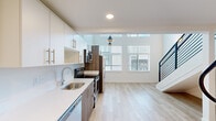 Located in Portland's vibrant West End neighborhood at 1670 SW Alder St, this modern loft-style unit showcases striking double-height ceilings that create an impressive sense of vertical space and openness. The main level features an open-concept layout where the kitchen flows seamlessly into the living area. The kitchen is outfitted with white upper cabinetry, warm wood-toned lower cabinets, stainless steel appliances including a refrigerator, electric range, microwave, and dishwasher, along with a clean white quartz countertop and an undermount sink. Light hardwood-style flooring runs throughout the main level, complementing the bright, white walls. Large windows flood the space with natural light and offer views of the surrounding streetscape. A sleek black metal staircase with horizontal railings leads to the upper loft level, which features plush carpet flooring, a ceiling fan, and additional windows that continue to bring in ample daylight. This loft area overlooks the main living space below through a matching open metal railing. The bathroom includes a walk-in shower with chrome fixtures. Recessed and pendant lighting throughout adds a polished, contemporary finish to this efficiently designed urban retreat.