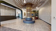Located at 5720 Peachtree Pkwy in Peachtree Corners, Georgia, this thoughtfully designed single-floor amenity space offers a generously sized, multi-functional environment that blends striking architectural details with curated social areas. The centerpiece of the main hall is a towering rope-wrapped column adorned with gold banding, from which decorative rope tendrils radiate outward across the ceiling, creating an organic, sculptural canopy above geometric-patterned tile flooring. A circular upholstered seating bench surrounds the column's base, inviting casual gathering.

Adjacent to the central hall, a lounge and library nook features floor-to-ceiling dark green built-in bookshelves flanking a wall-mounted television, textured accent wallcovering, and plush burnt-orange velvet armchairs set atop a richly patterned area rug. Original artwork accents the walls, adding warmth and character.

The catering kitchen is a standout feature, lined on both sides with slate-blue cabinetry, white quartz countertops, a turquoise glass tile backsplash, a wine refrigerator, and dishwasher. Large windows flood the space with natural light, while slatted wood ceiling panels and a statement chandelier define the zone. A billiards table anchors the game area beyond. A wood slat partition screen elegantly separates zones, maintaining visual openness throughout.