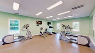 Located at 1400 Boxwood Blvd in Columbus, Georgia, this fitness center offers a comprehensive and thoughtfully organized workout facility spread across a single floor. The space is finished with soft mint-green walls and durable rubber flooring, creating an energizing yet comfortable environment for exercise.

The cardio area is well-stocked with treadmills, elliptical trainers, and a stationary bike, all arranged to allow ample space between equipment. Wall-mounted flat-screen televisions provide entertainment during workouts. The strength and functional training section features a cable machine system, an adjustable weight bench, kettlebells, medicine balls, and a colorful array of stability and exercise balls neatly organized on dedicated racks.

A large wall mirror in the main workout area enhances the sense of space and allows users to monitor their form during exercise. The facility benefits from generous natural light streaming through multiple white-framed windows, which also offer views of the surrounding outdoor area. Recessed panel lighting ensures the space remains well-lit throughout.

The layout is open and spacious, accommodating both cardio and strength training without feeling crowded. The clean, functional design and variety of equipment make this fitness center a versatile resource for a broad range of workout routines and fitness goals.