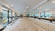 Located at 1888 Brittmoore Rd in Houston, Texas, this contemporary fitness studio is a well-appointed exercise space designed to support a variety of workout activities. The studio features a clean, modern aesthetic with crisp white walls, recessed ceiling lighting, and patterned low-pile carpeting that spans the entire floor area.

A defining feature of the room is the expansive floor-to-ceiling mirror wall, which runs the full length of one side and is accompanied by a wall-mounted ballet barre — ideal for stretching, barre classes, or dance-style workouts. This mirrored wall effectively amplifies the sense of space and enhances the bright, open atmosphere of the studio.

Large black-framed floor-to-ceiling windows line the opposite wall, flooding the interior with abundant natural light and offering views of an outdoor pool and landscaped surroundings. Spinning bikes and medicine balls are among the fitness equipment visible within the space, making it well-suited for cycling and cross-training sessions.

Access to the studio is provided through sleek black-framed glass sliding doors, which connect the space to an adjacent lounge or common area, as well as a private door that leads further into the facility. A ceiling-mounted audio unit supports group fitness classes. The overall design balances functionality with a polished, contemporary finish.